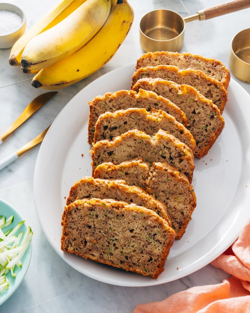 Banana zucchini bread sliced on a plate and surrounded by ingredients and measuring cups